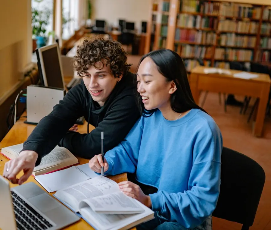 Two students working through a textbook together on a laptop in a library